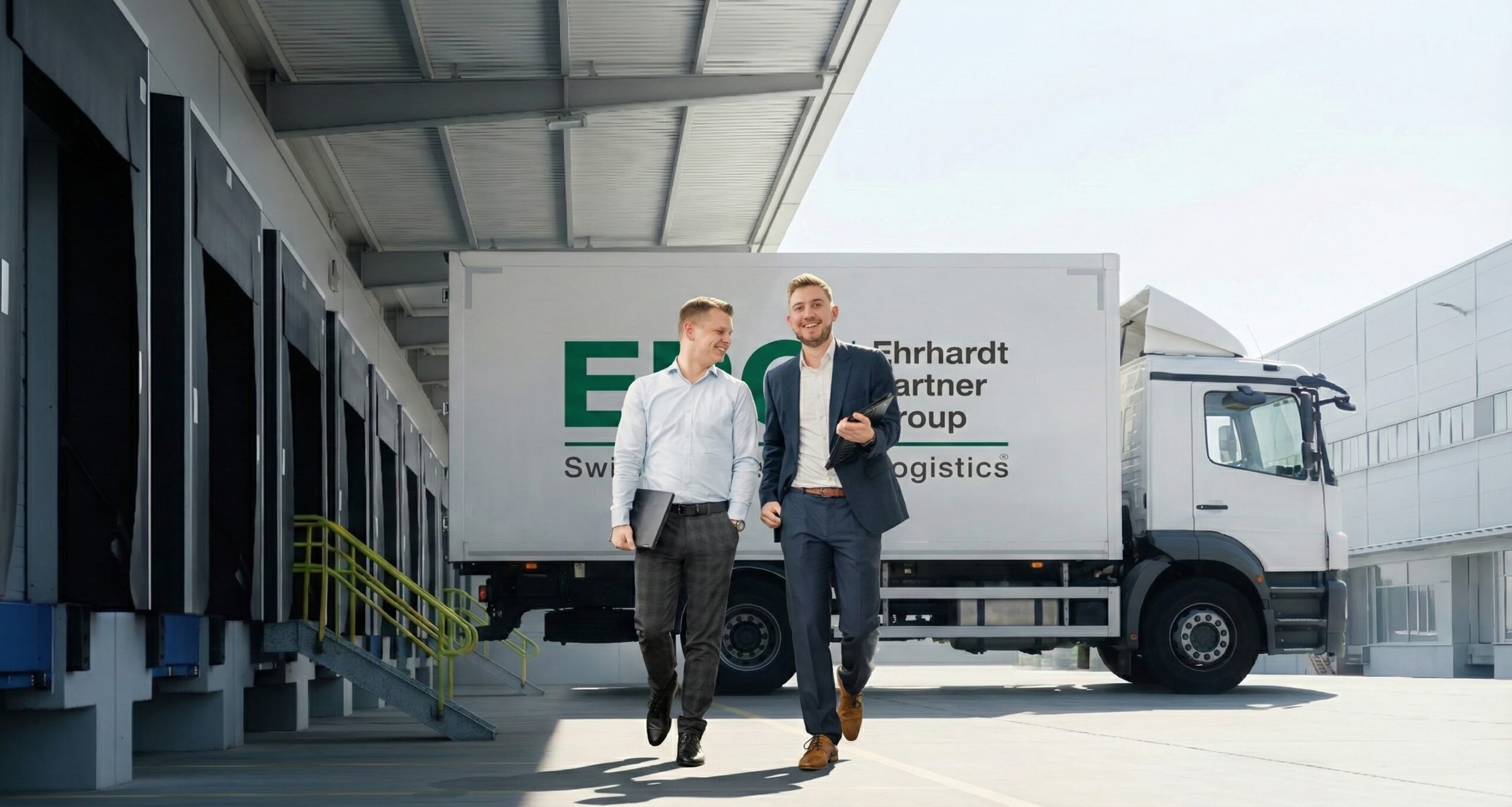 Zwei Männer vor einem Logistik-Lkw, Lagerhaus im Hintergrund.